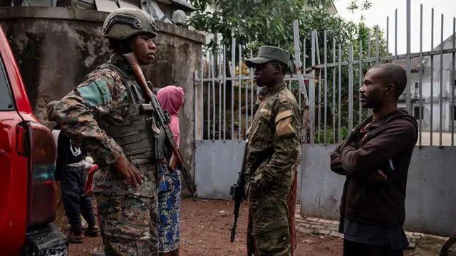 Deux militaires en uniforme, fusils à la main, accompagnés d’un civil et d’une femme en arrière-plan